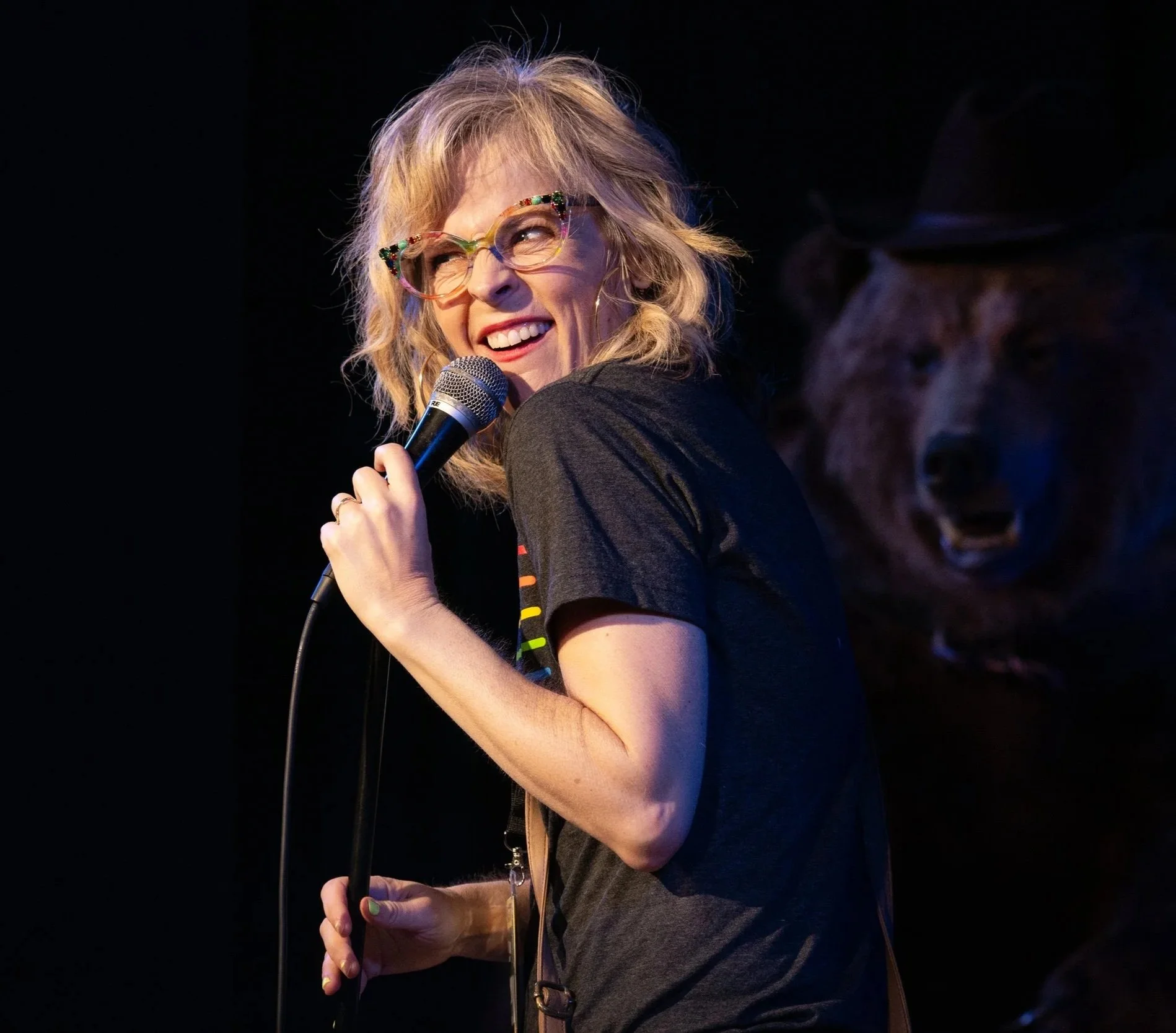 A woman with blonde curly hair and colorful glasses smiling and holding a microphone on stage with a dark background and a blurry person in the background.