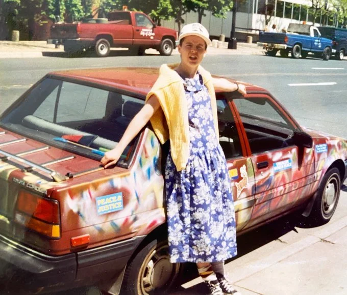 A young woman standing next to a colorful spray-painted car on a city street, wearing a blue floral dress, a white hat, and a yellow towel draped over her shoulder.
