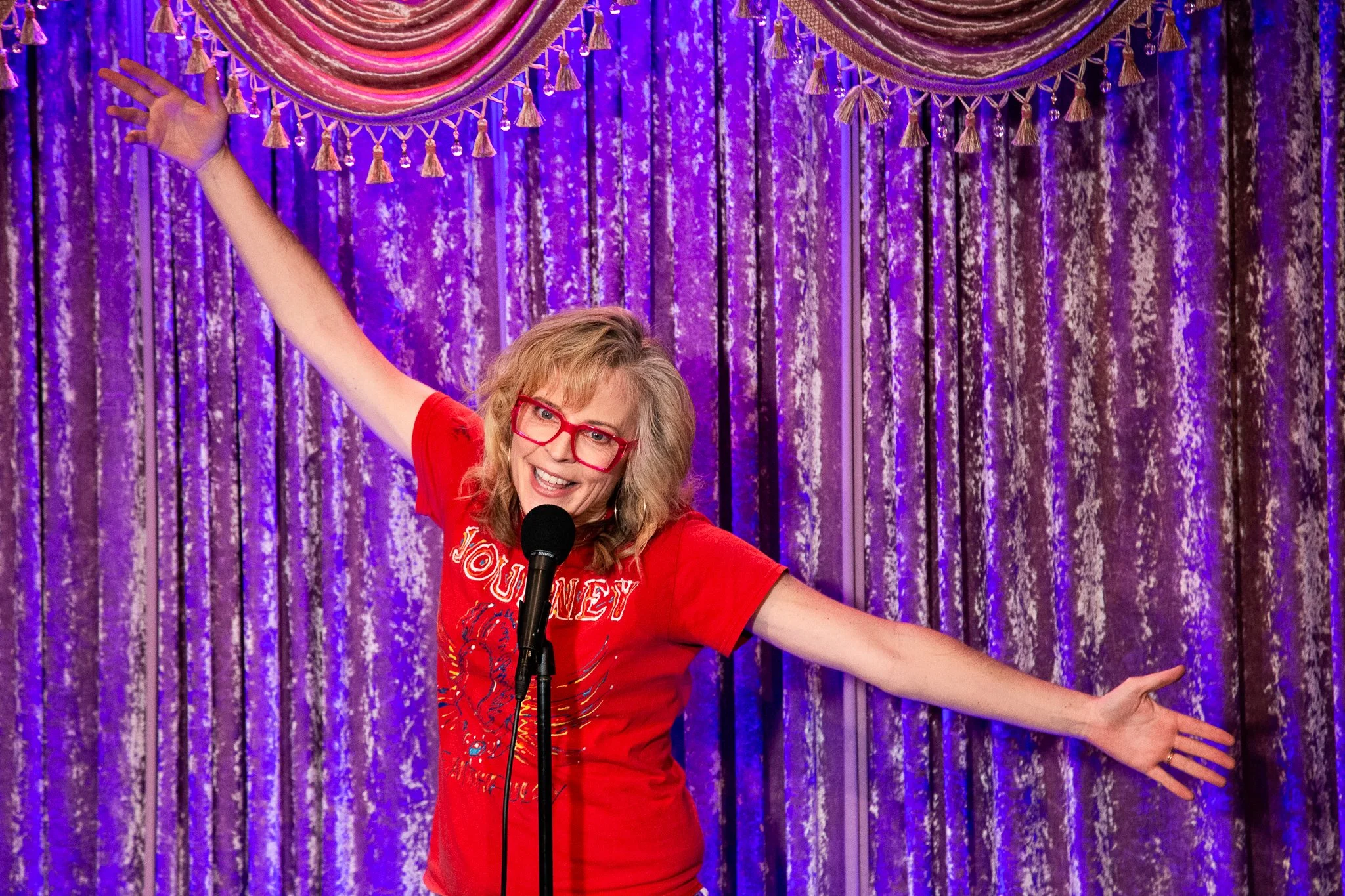 Woman with curly blonde hair and red glasses smiling, wearing a red shirt with text, standing in front of purple velvet curtains, holding a microphone in front of her mouth, with arms outstretched.