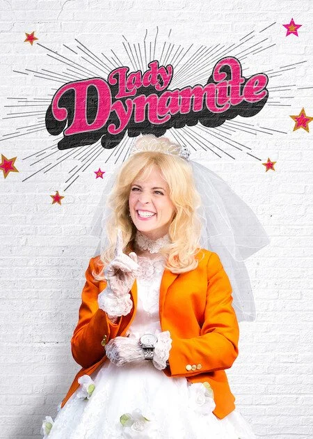 A woman dressed in a wedding gown with an orange blazer, wearing a blonde wig and white veil, standing in front of a wall with a sign that reads 'Lady Dynamite' surrounded by stars.
