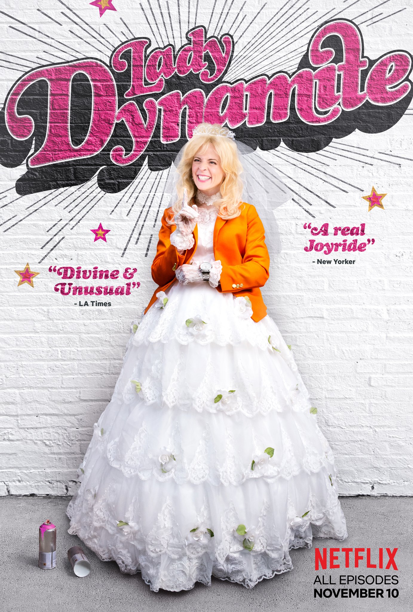 A woman dressed in a wedding gown and an orange blazer, smiling and posing in front of a wall with promotional text and graphics. There are spray paint cans on the ground beside her.