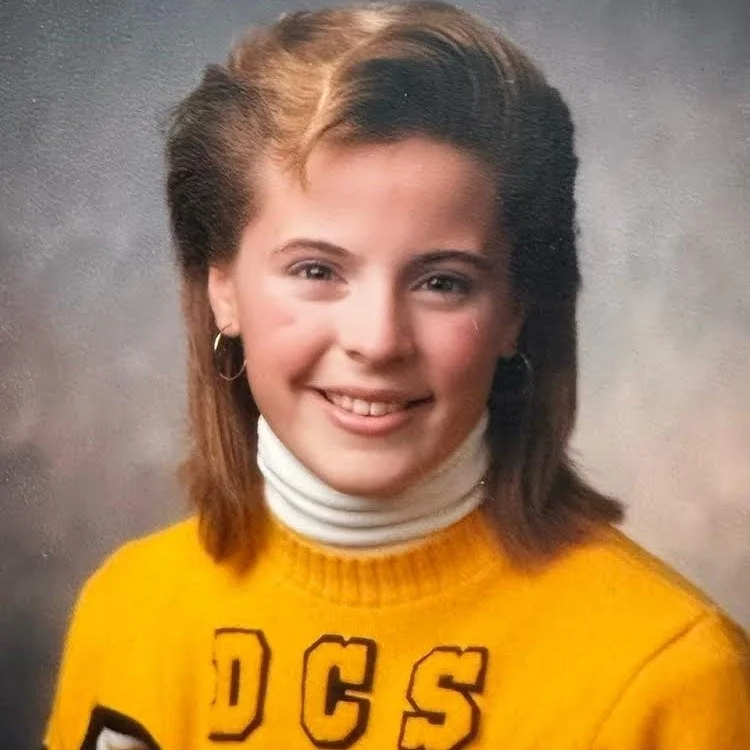 A young woman with shoulder-length brown hair, wearing a yellow sweatshirt with the letters 'DCS' on it, and a white turtleneck underneath, smiling at the camera.