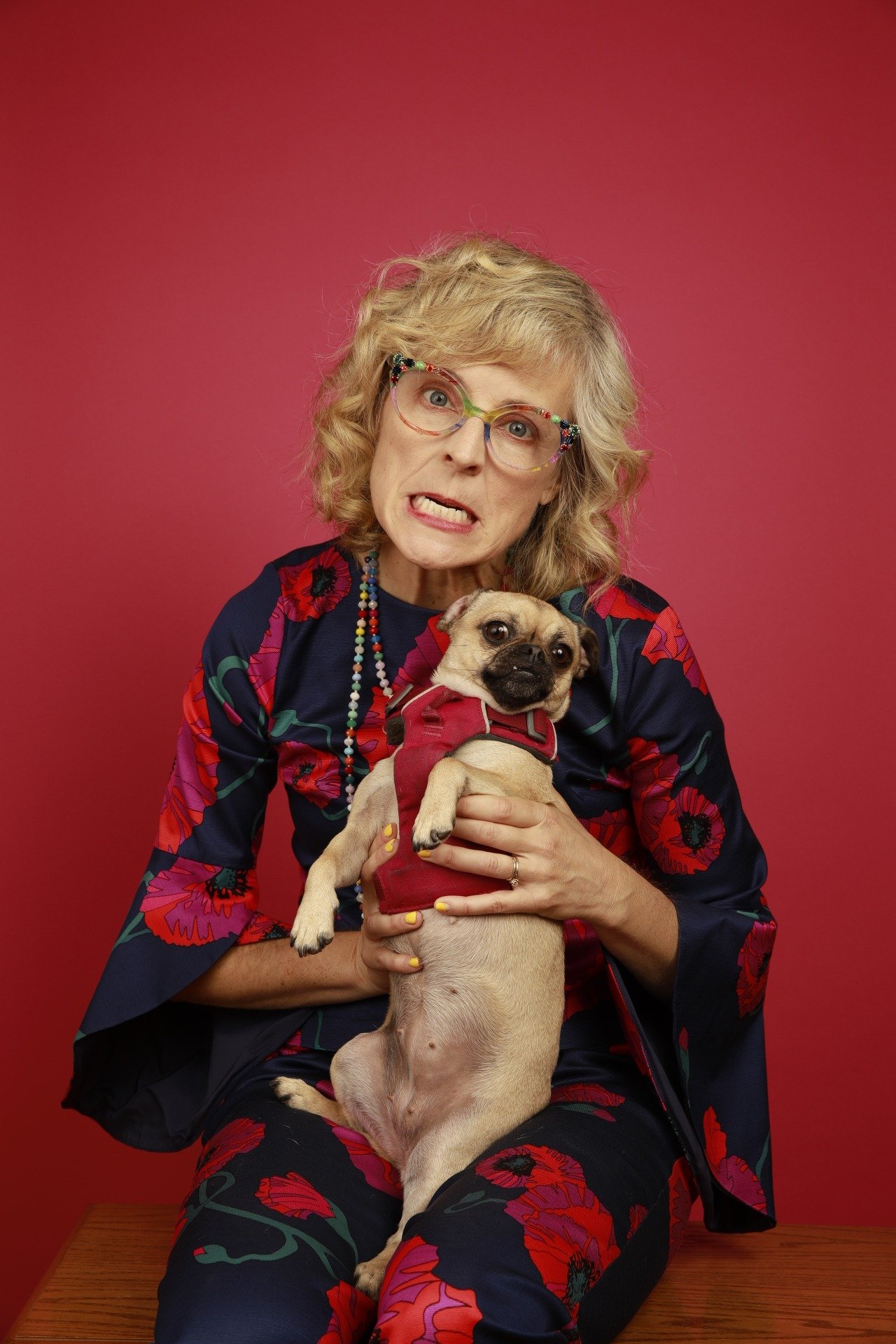 A woman with curly blonde hair, colorful glasses, and floral clothing holding a small pug puppy in front of a red background.