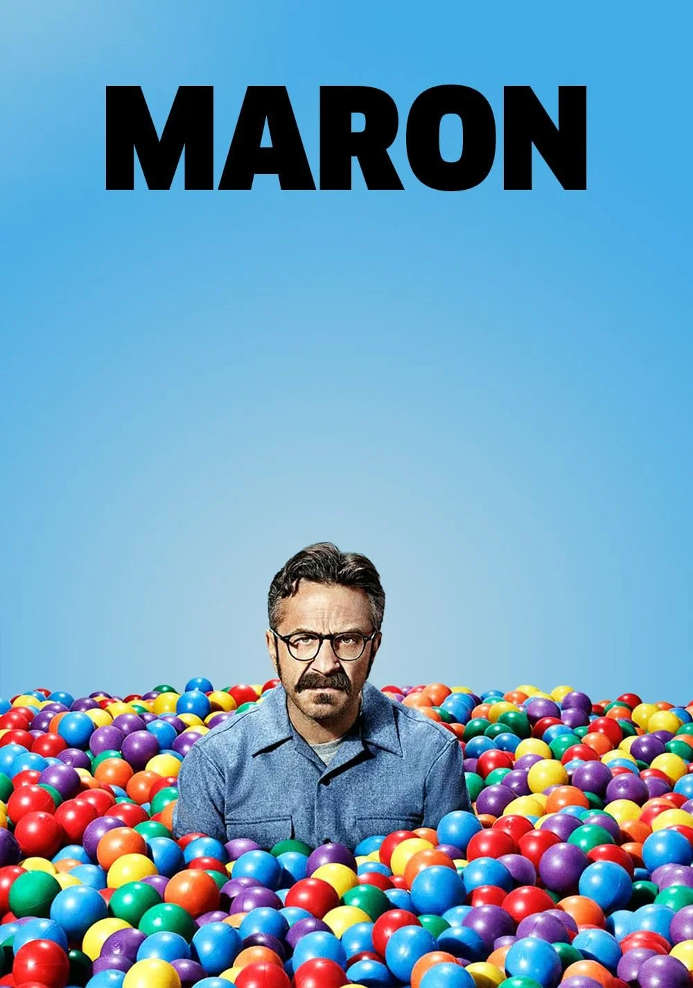 A man with glasses and a mustache sitting in a colorful ball pit with a blue sky background. The top of the image has the word "MARON" in bold black letters.