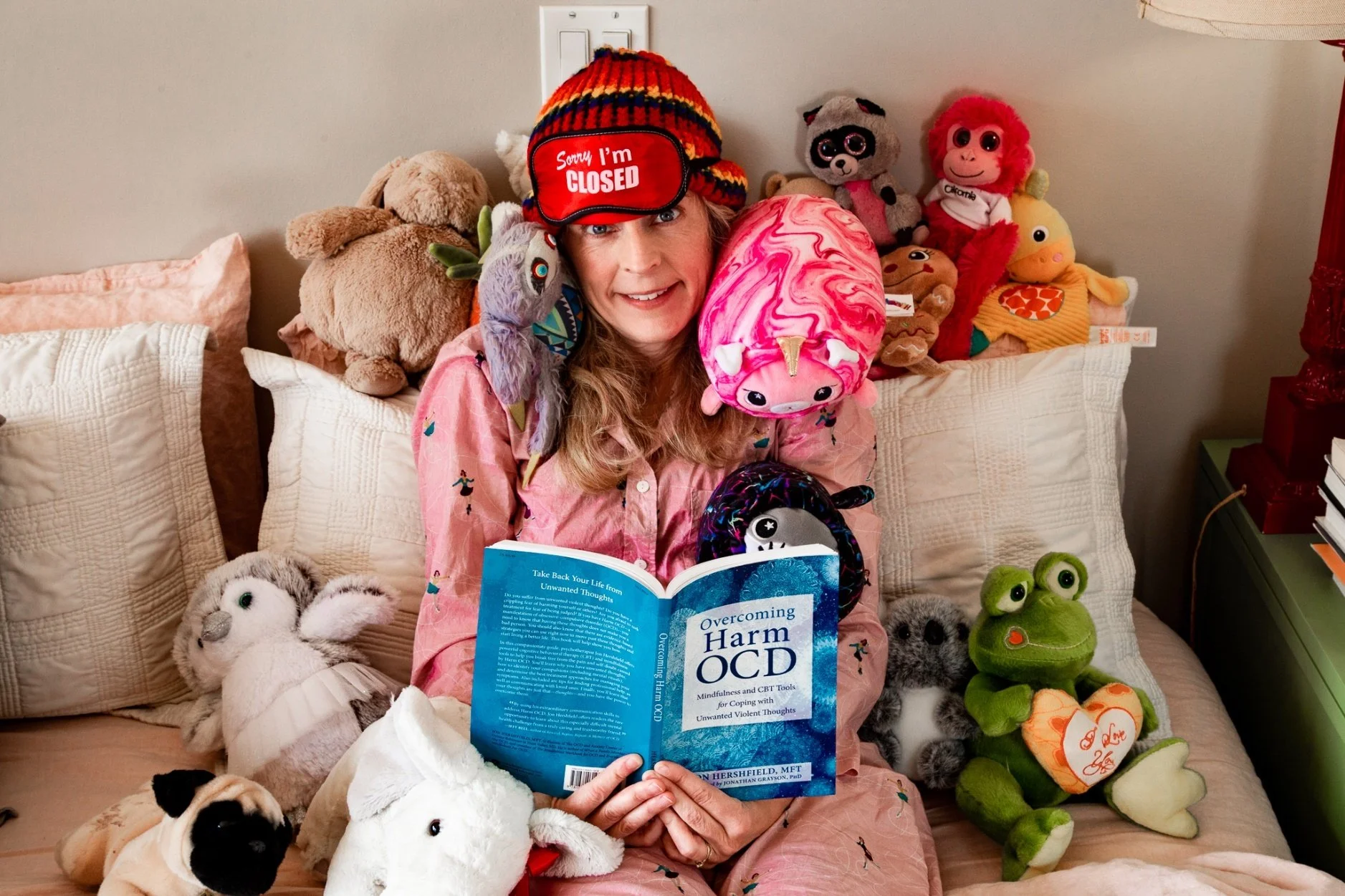A woman sitting on a beige couch surrounded by various stuffed animals, holding an open book titled 'Overcoming Harm OCD'. She wears a colorful striped hat with a sign that says 'Sorry I'm CLOSED' and has a cheerful expression.