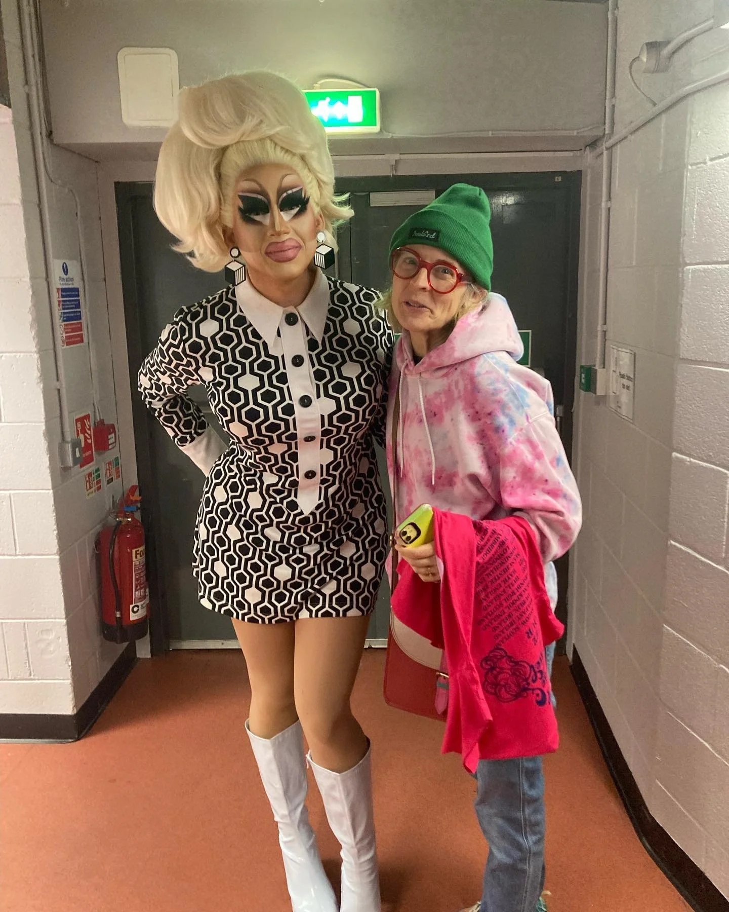 A person in elaborate drag makeup and outfit stands next to a woman in casual clothing, wearing a green beanie and red glasses, in a hallway with white cinder block walls, a fire extinguisher, and exit sign visible.
