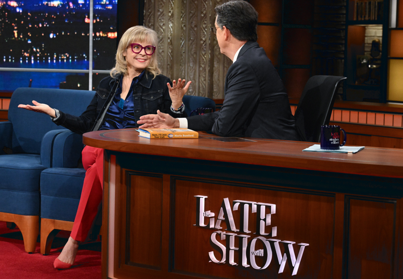 A woman in red pants and a dark jacket is talking with a man in a suit during a talk show. They are seated at a wooden desk with the sign 'the LATE SHOW' and a mug. The woman is smiling and gesturing with her hands, while the man is listening.