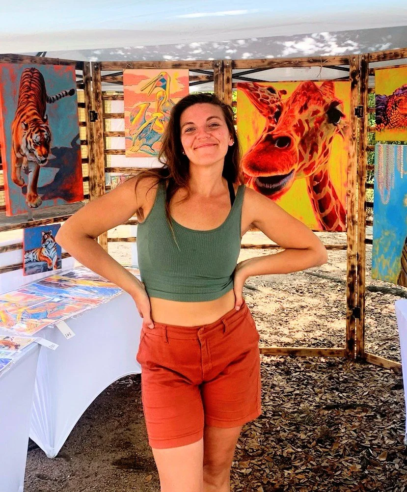 A woman standing in front of colorful animal paintings at an outdoor art market, smiling with her hands on her hips.