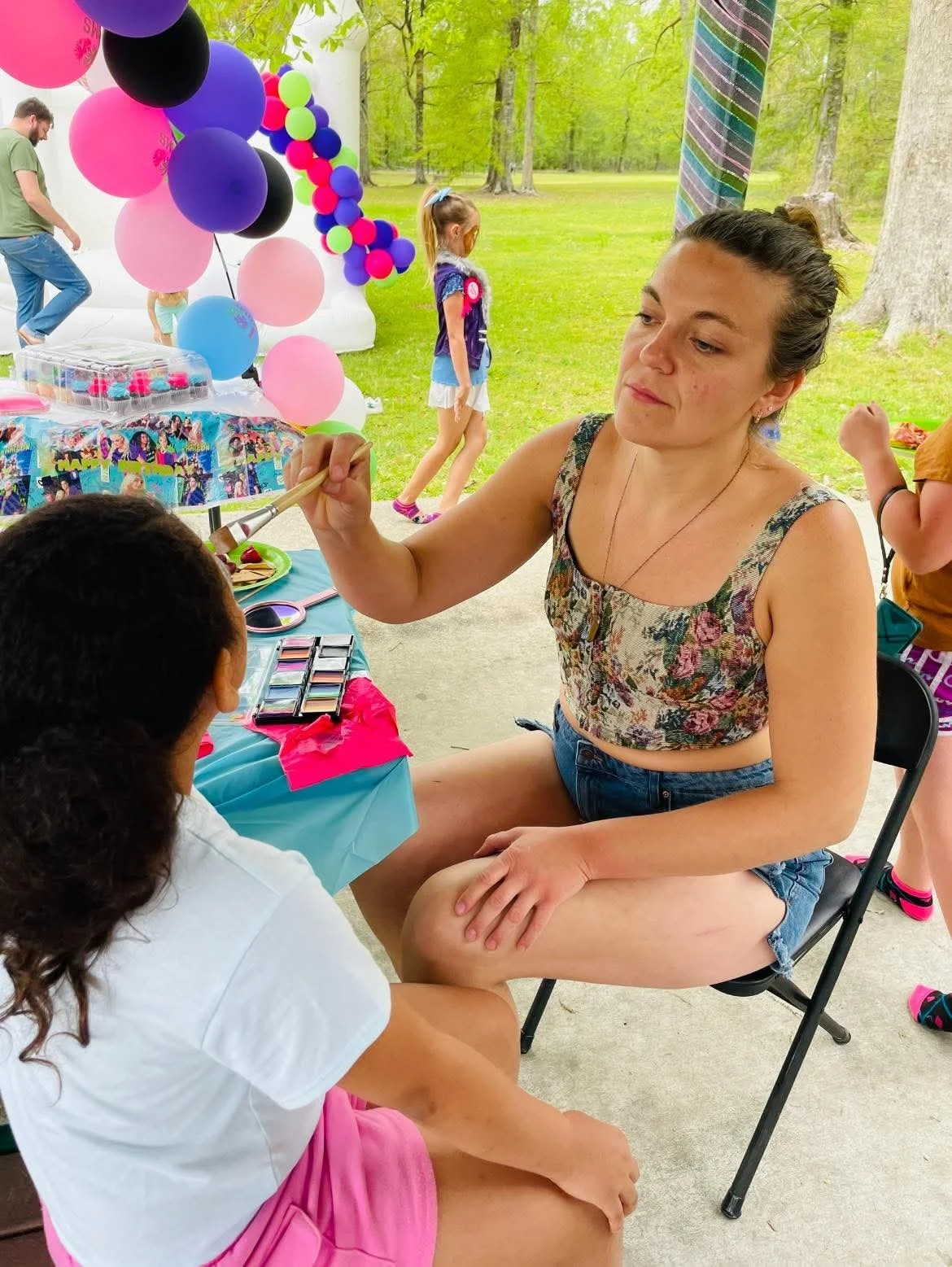 Woman painting face of a girl at an outdoor birthday party with balloons and kids in the background.