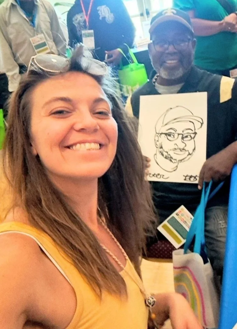 A smiling woman taking a selfie at a busy event, with people holding a caricature drawing of a man wearing glasses and a cap in the background.