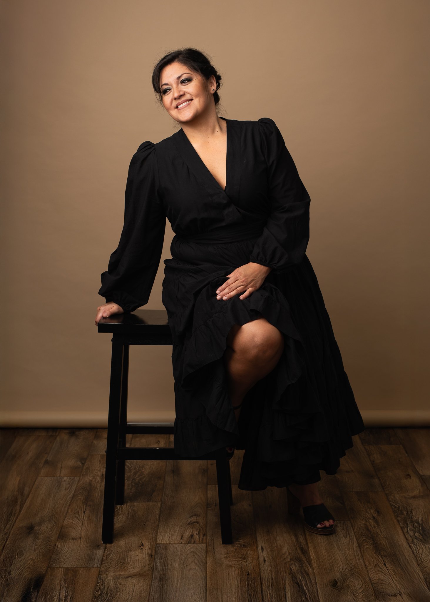 Woman in a long black dress sits on a wooden stool, smiling, with one hand on her knee, against a brown background.