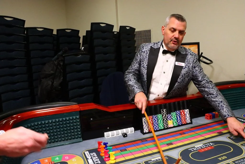 A casino dealer in a patterned tuxedo oversees a table game, using a stick to move chips and dice on the gaming table.