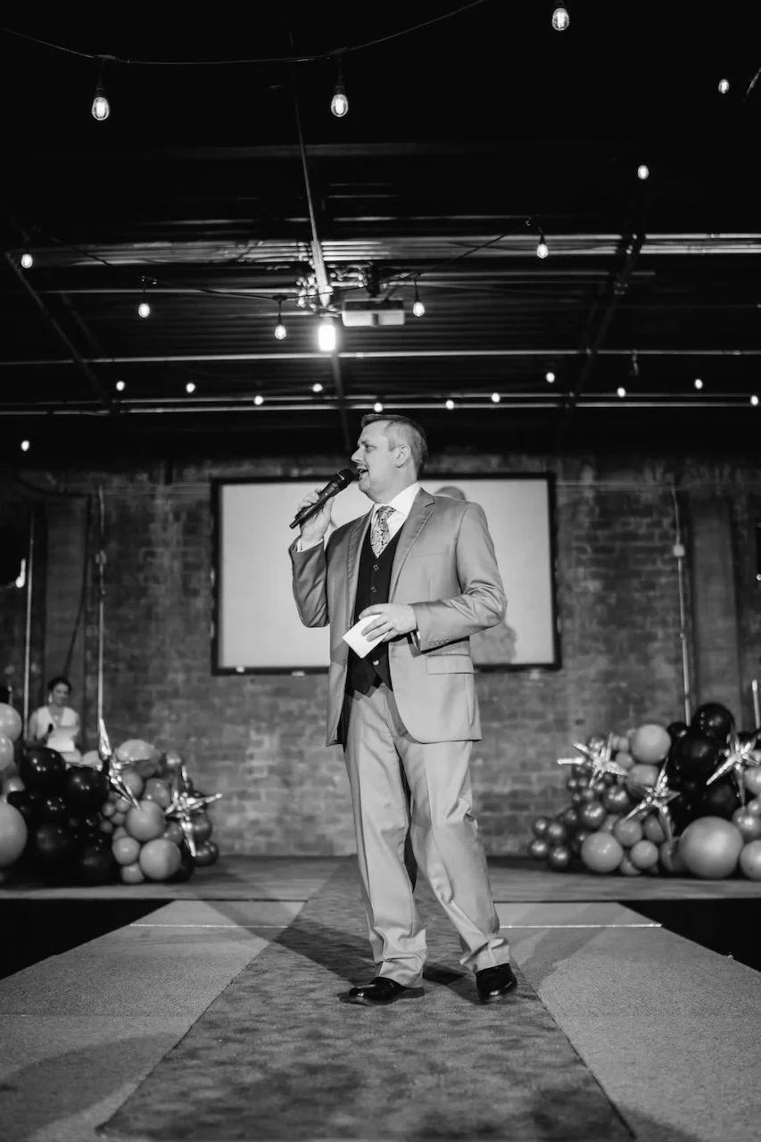 Man in a suit holding a microphone speaks on stage with balloons and string lights in the background. Black and white image.
