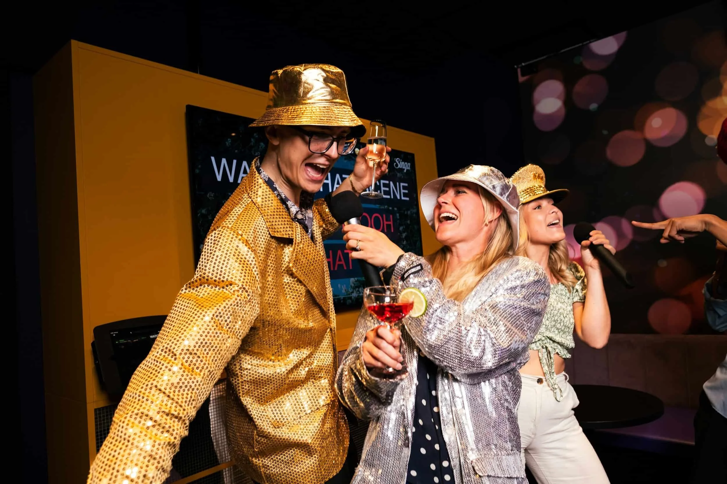 Three people in shiny jackets and hats sing and laugh at karaoke, holding drinks and a microphone, with colorful lights in the background.