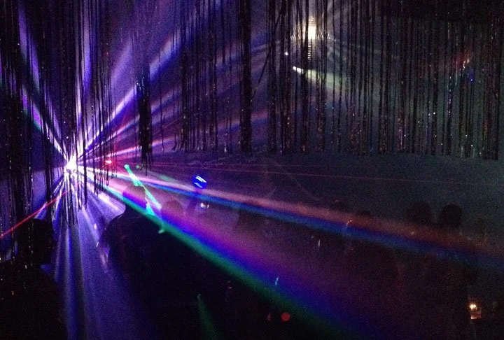 Silhouettes of people dancing with colorful laser lights and beams shining in a dark, smoky party atmosphere.