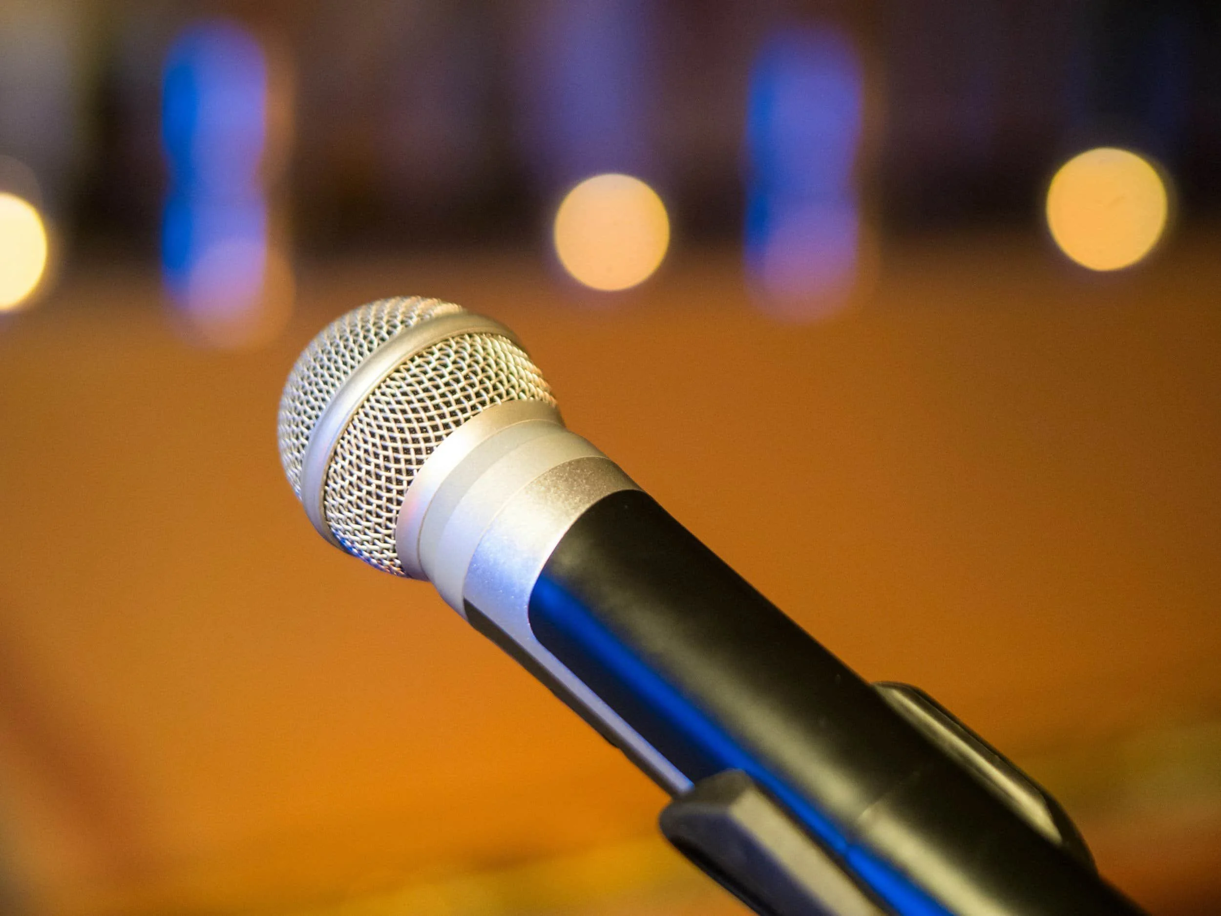 Close-up of a silver microphone on a stand with a blurred, warm-colored background and bokeh lights.