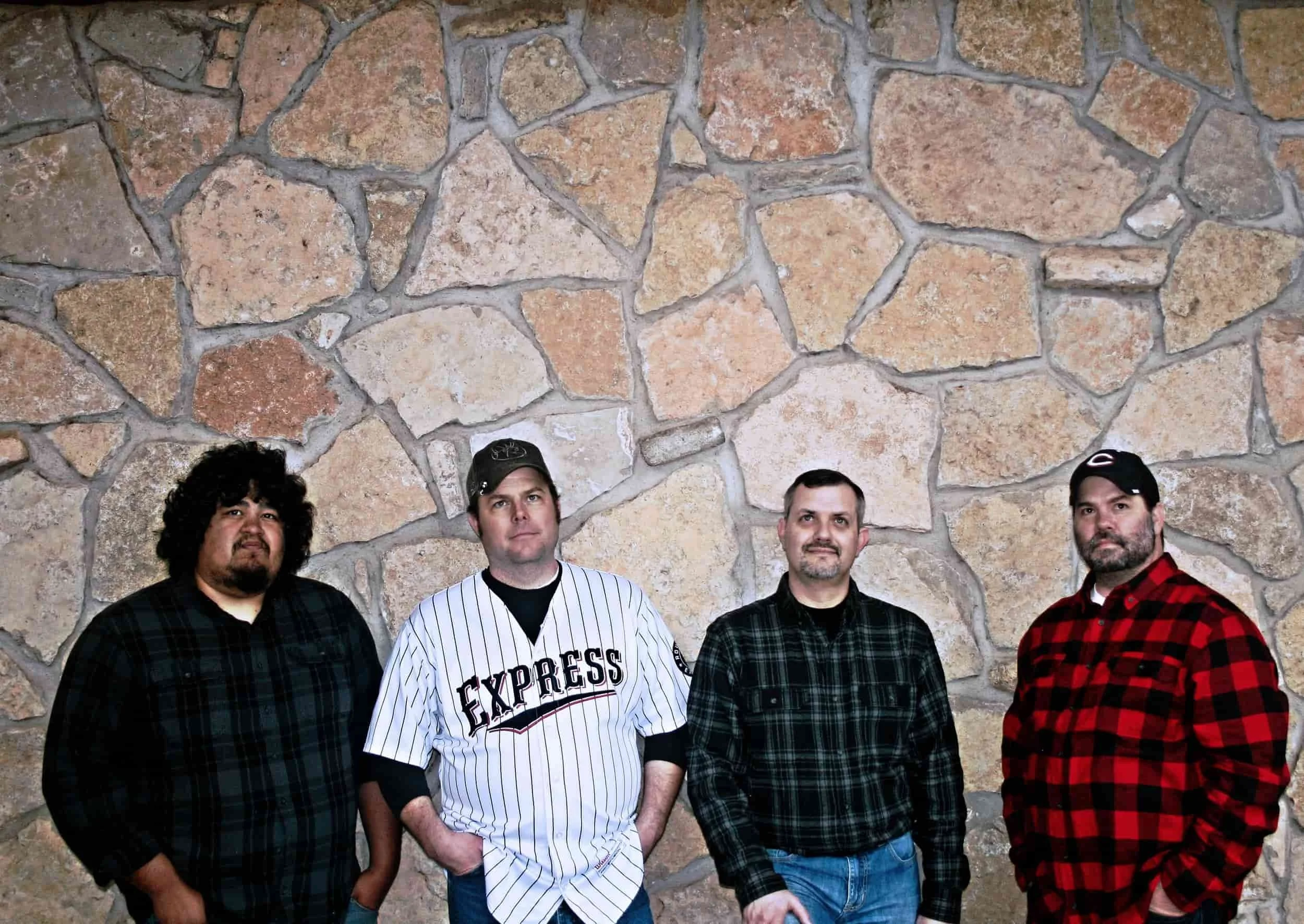 Four men stand in front of a stone wall, dressed in casual shirts and jackets, facing the camera with neutral expressions.