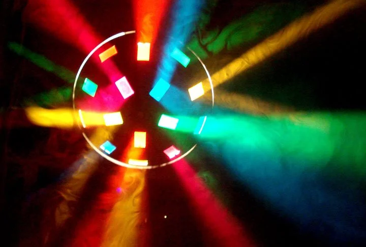Colorful beams of red, yellow, blue, and green light radiate from a circular disco light in a dark setting.