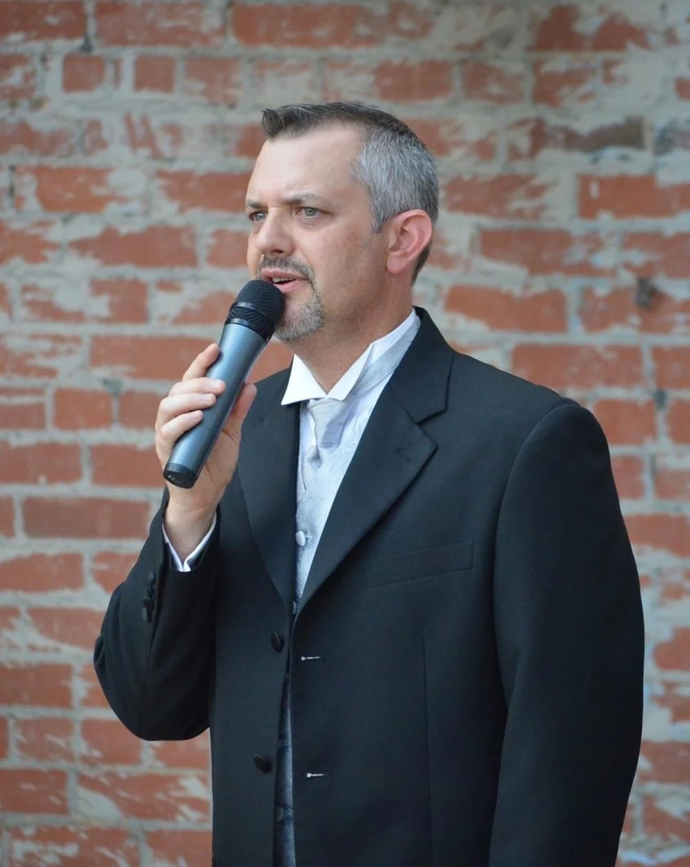A man in a suit holds a microphone and speaks in front of a red brick wall.