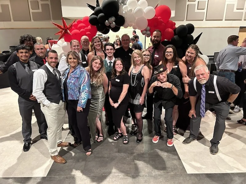 A group of people dressed up, smiling and posing under a red, black, and white balloon arch at an indoor event or party.