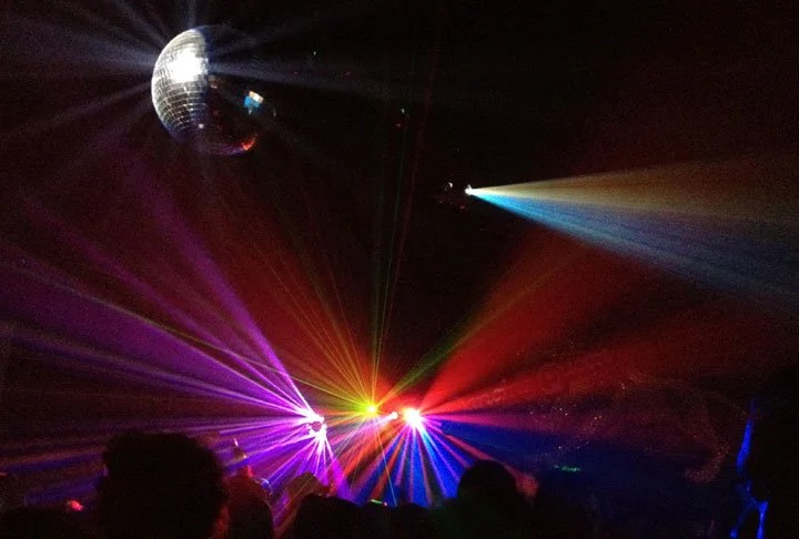 A disco ball and colorful stage lights create vibrant beams over a dark, crowded dance floor.