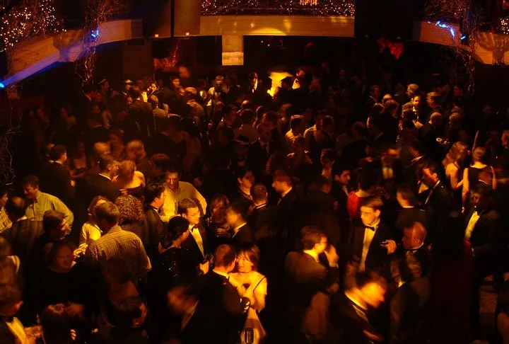 A crowded ballroom filled with people in formal attire, mingling and dancing under warm, dim lighting.