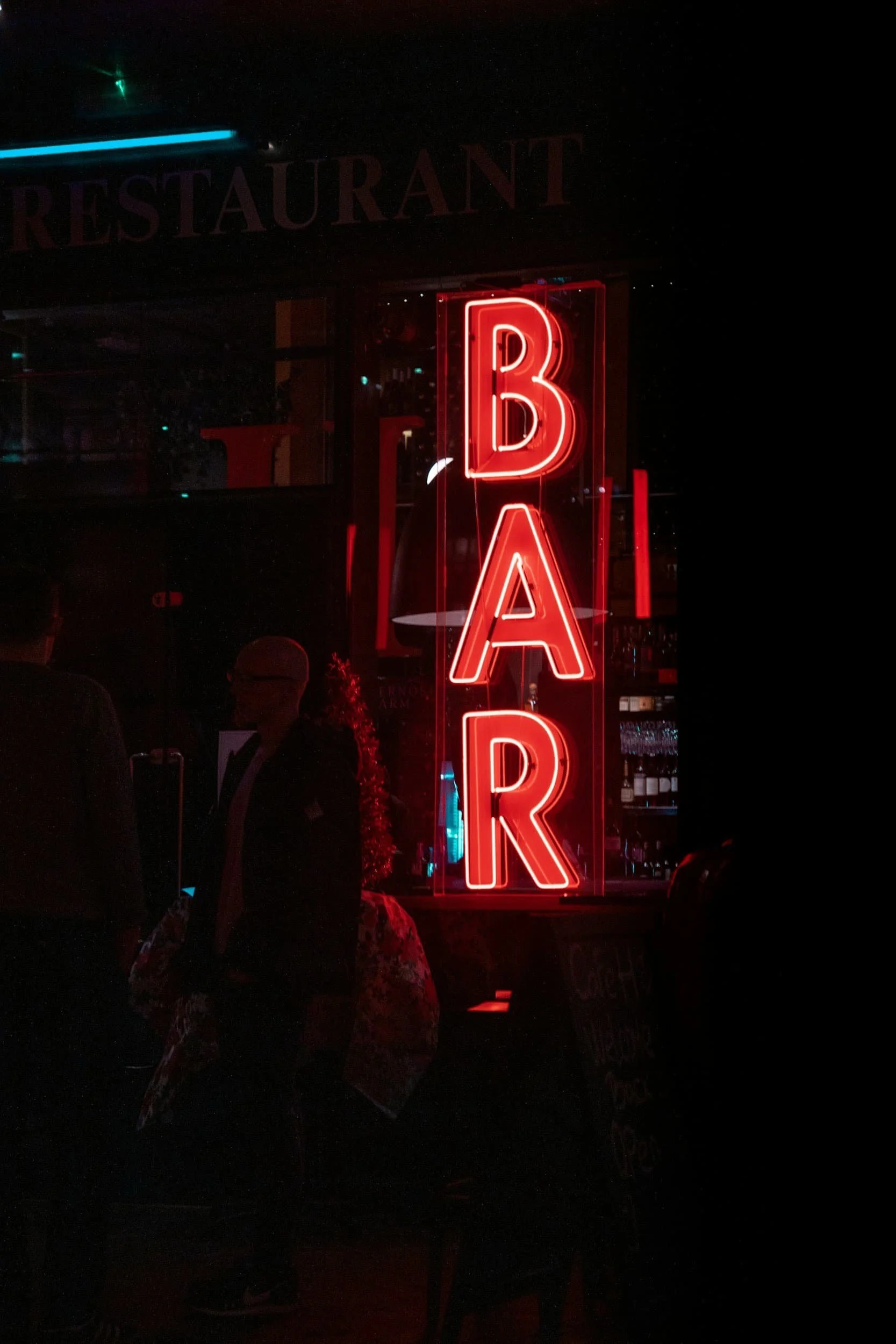 A bright red neon BAR sign glows at night, partially illuminating people standing outside a dark bar or restaurant.