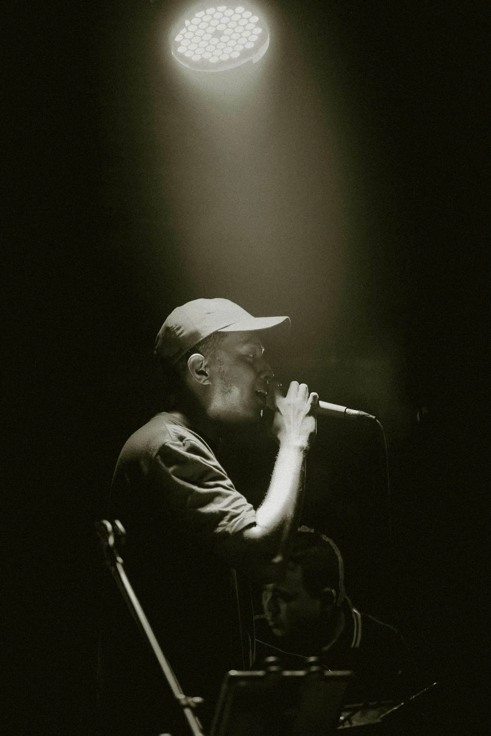 A man in a cap sings into a microphone under a spotlight on a dark stage, with another person visible in the background.
