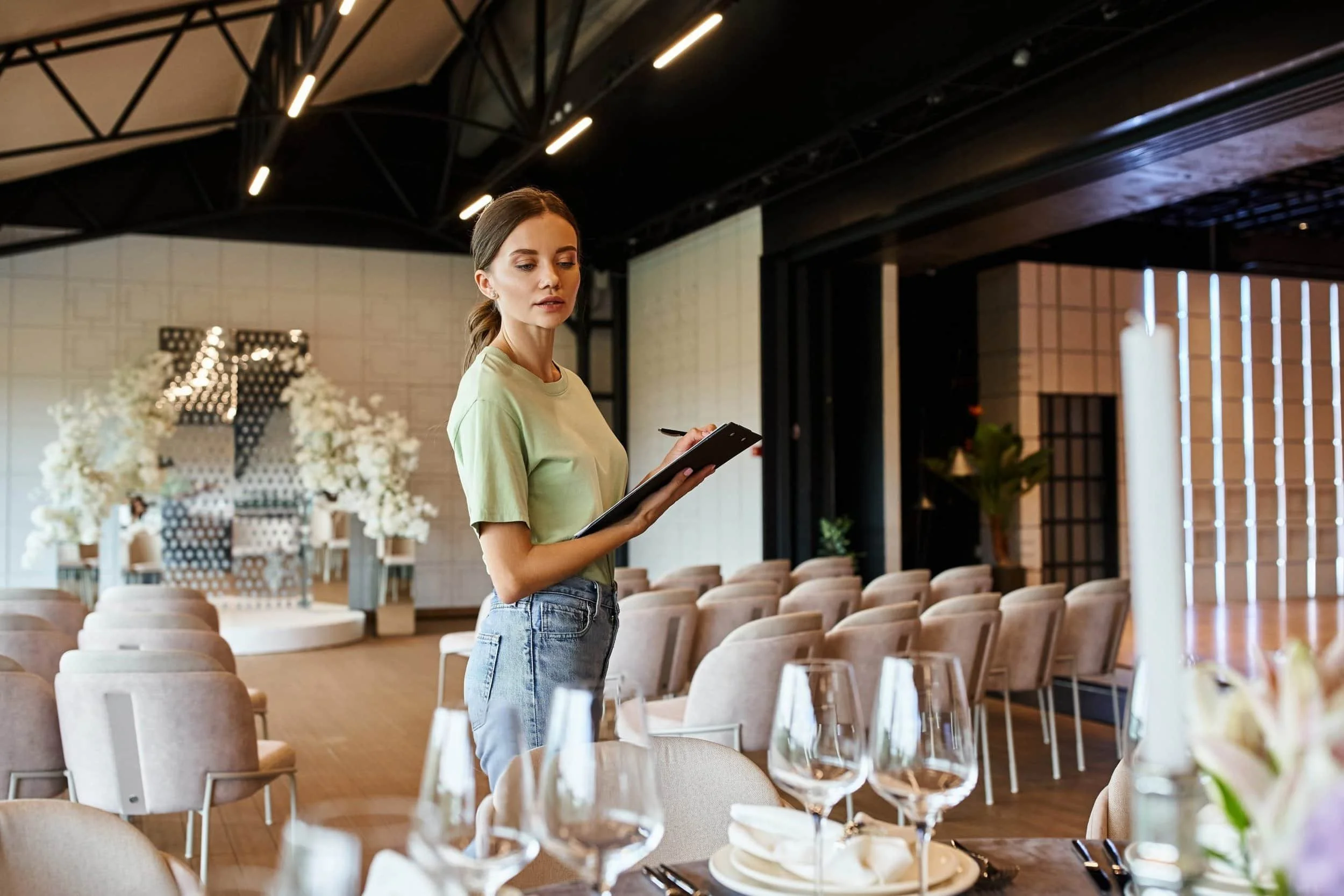 Woman with a clipboard checks arrangements in a decorated event hall set up with chairs, tables, and floral displays.