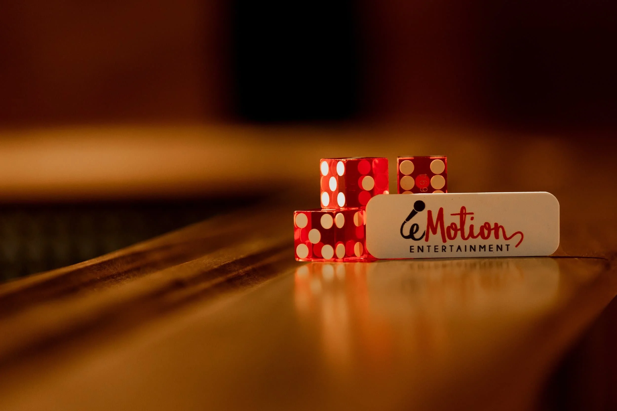 Three red dice stacked next to a white nametage labeled eMotion Entertainment on a wooden surface, blurred background 