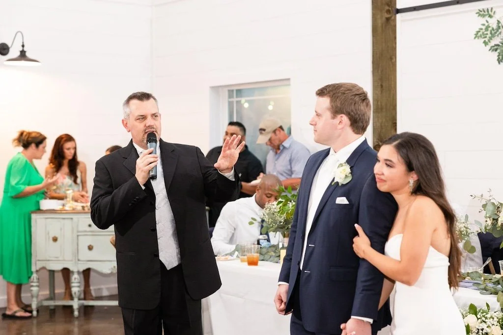 A man speaks into a microphone beside a bride and groom at a wedding reception, while guests are gathered in the background.