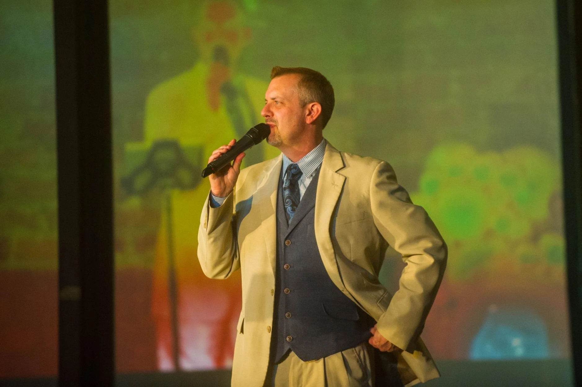 Man in a beige suit holds a microphone and speaks on stage with colorful, blurred background projection.