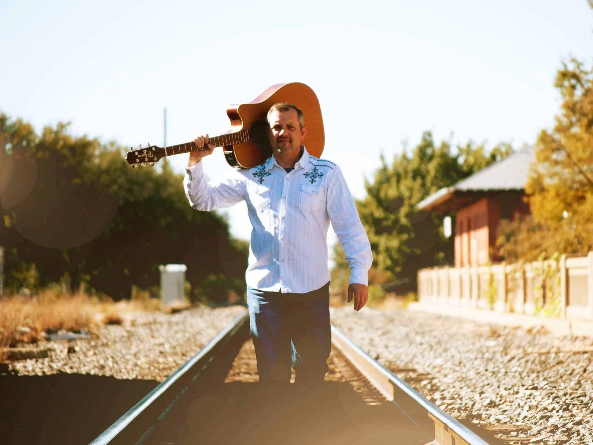 Man in a white shirt walks on train tracks carrying an acoustic guitar over his shoulder; trees and a building in the background.