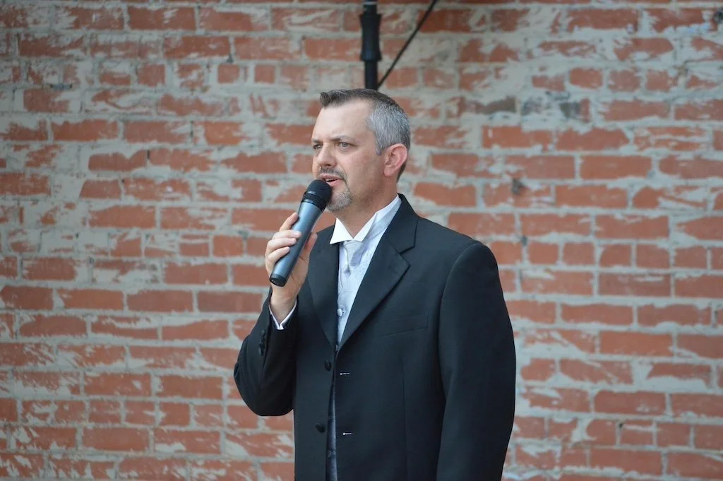 A man in a suit holds a microphone and speaks in front of a red brick wall.