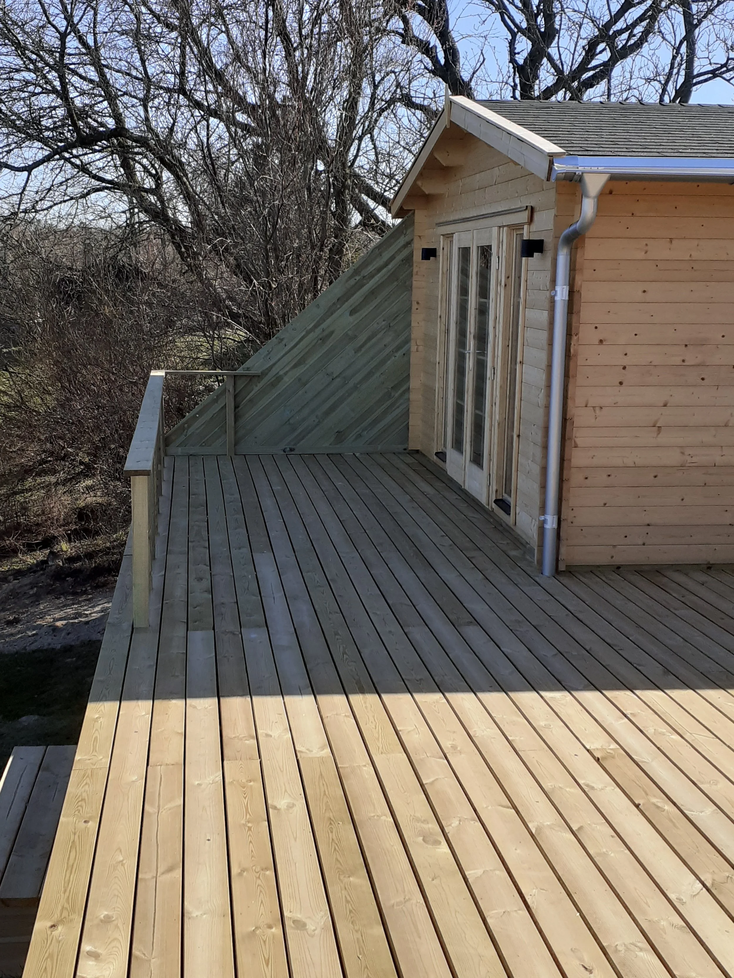 Træterrasse ved hus med skydedør og nyt træhegn, omgivet af natur og træer.