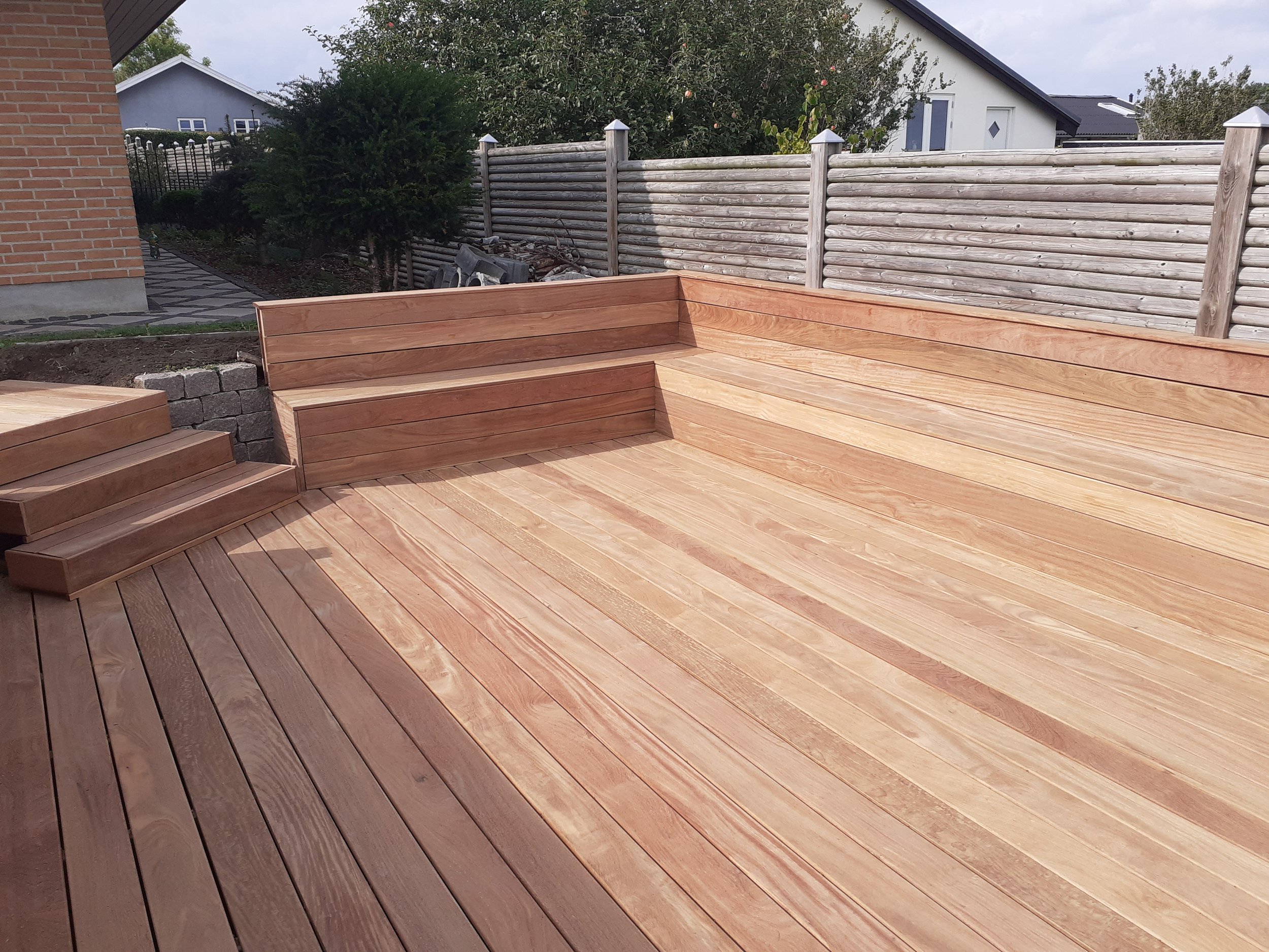 Ny træterrasse med trappe i baghave, omgivet af hegn og nabohuse.