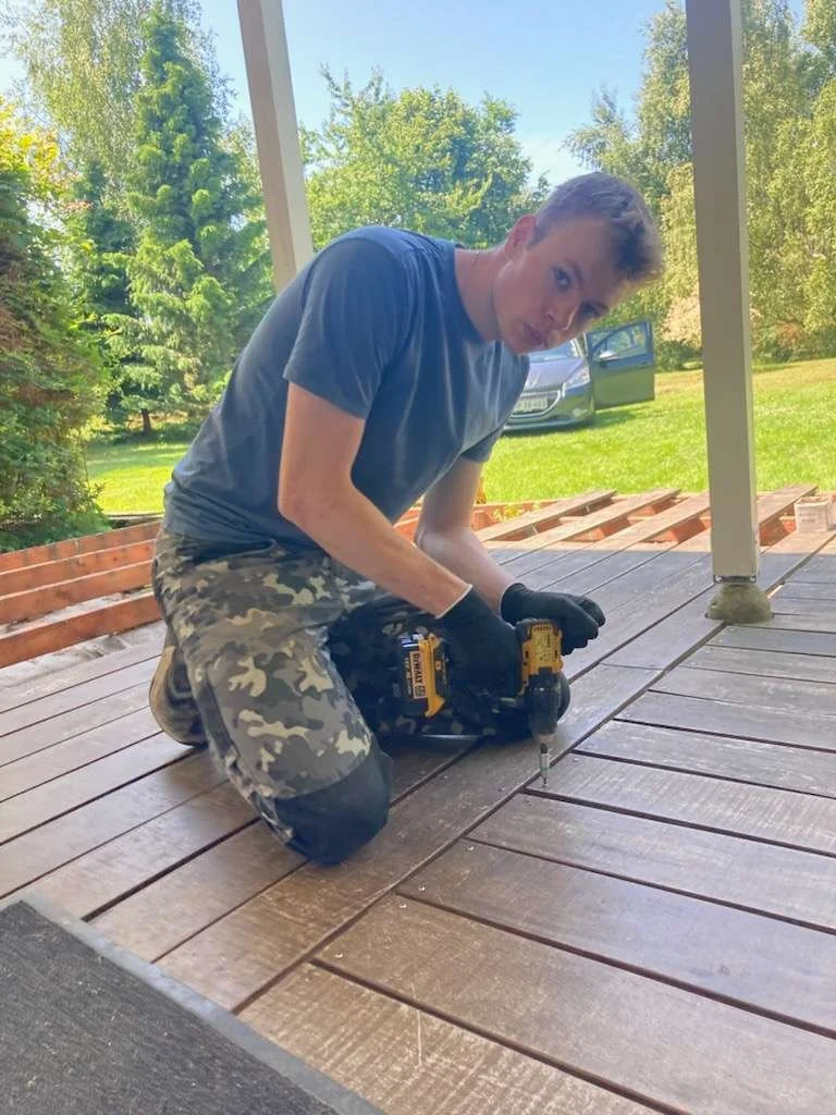 En ung håndværker arbejder på en træterrasse og monterer brædder med en skruemaskine.