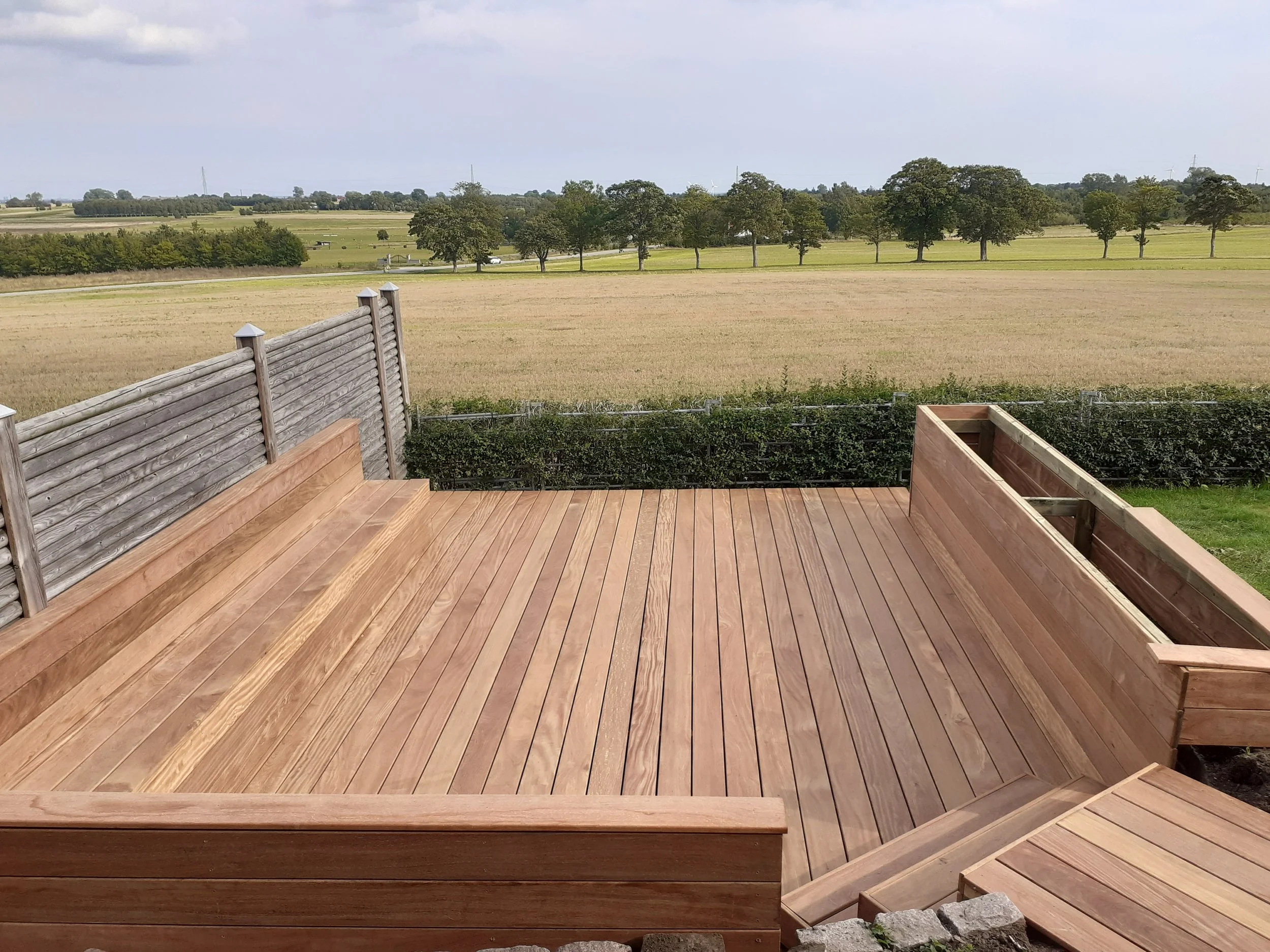 Træterrasse med udsigt over åbent landskab med marker og træer.