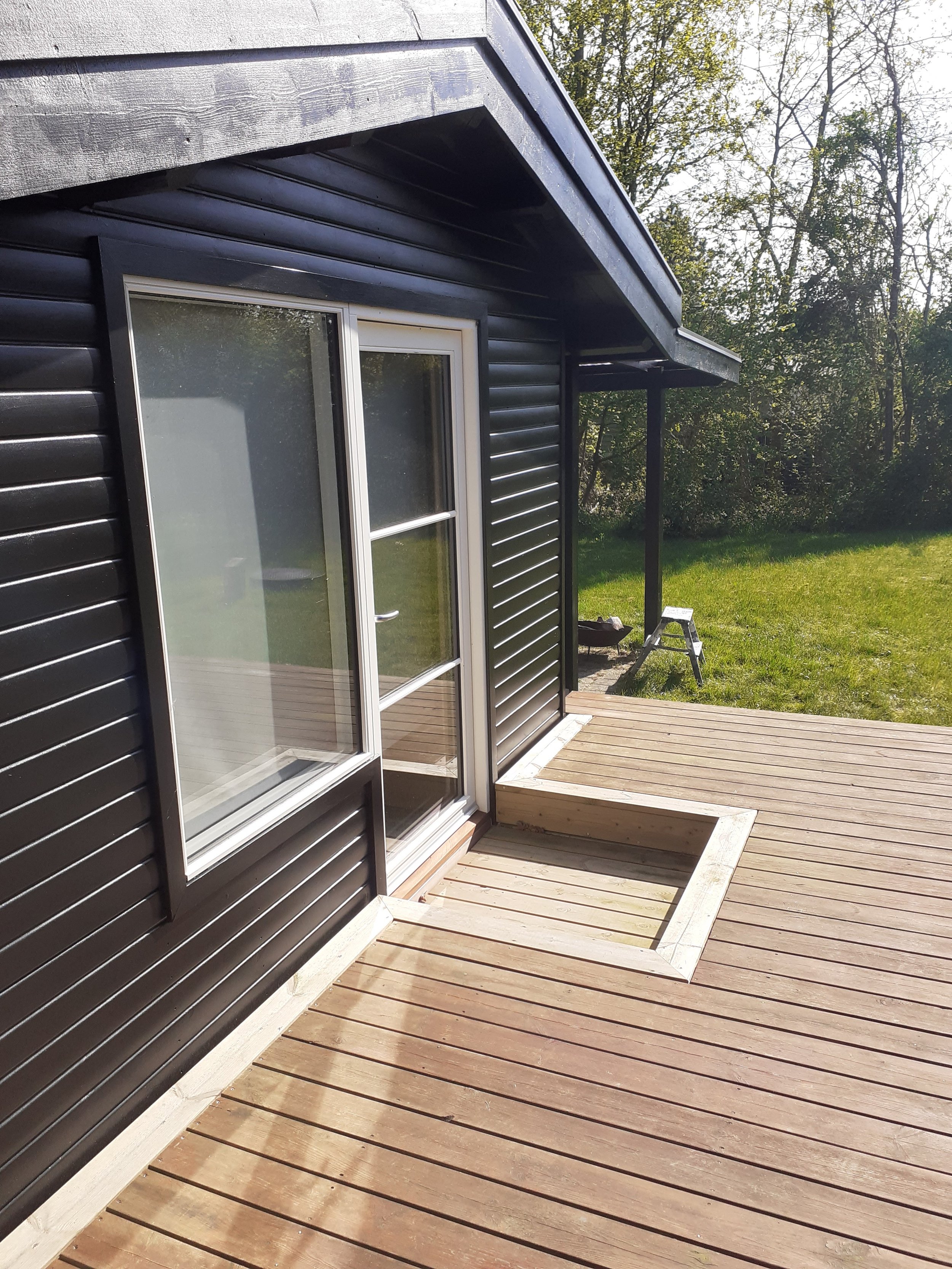 Træterrasse ved sort hus med skydedør i glas og indbygget område, egnet til fx spa eller plantekasser.