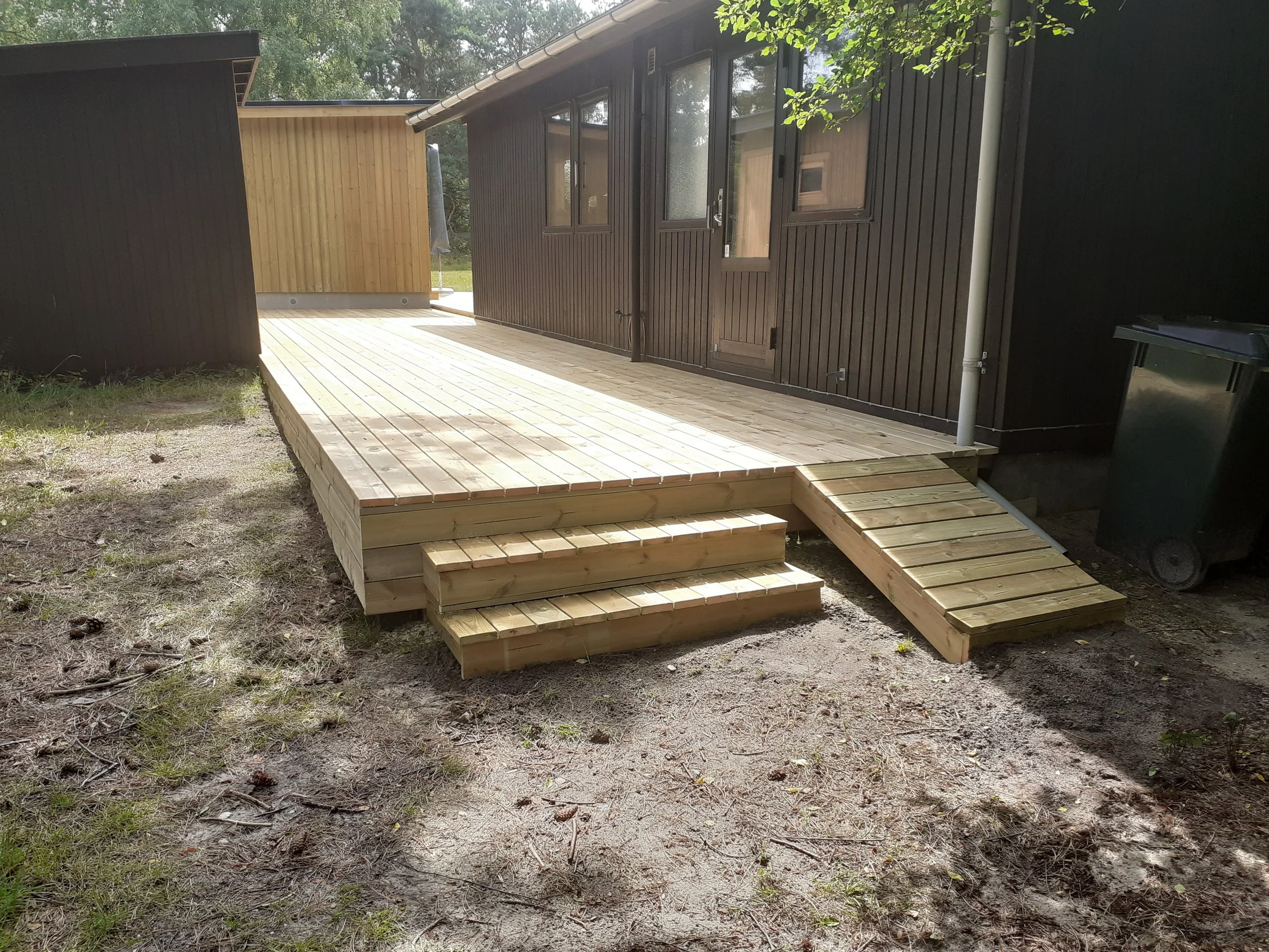 Træterrasse med trappe og rampe ved et mørkt hus, placeret i en baghave med græs og træer.