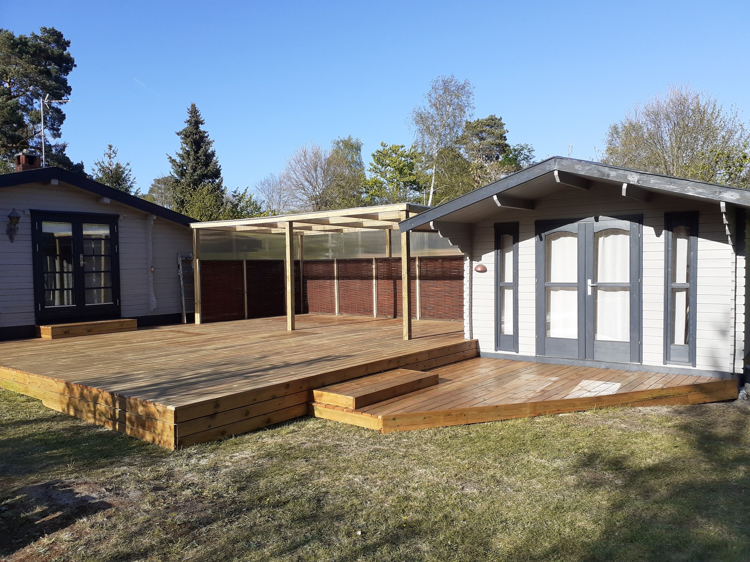 Baghave med nyopført træterrasse ved et mindre hus. Terrassen har trappe ned til haven samt et afskærmet område med gennemsigtige paneler og træhegn.