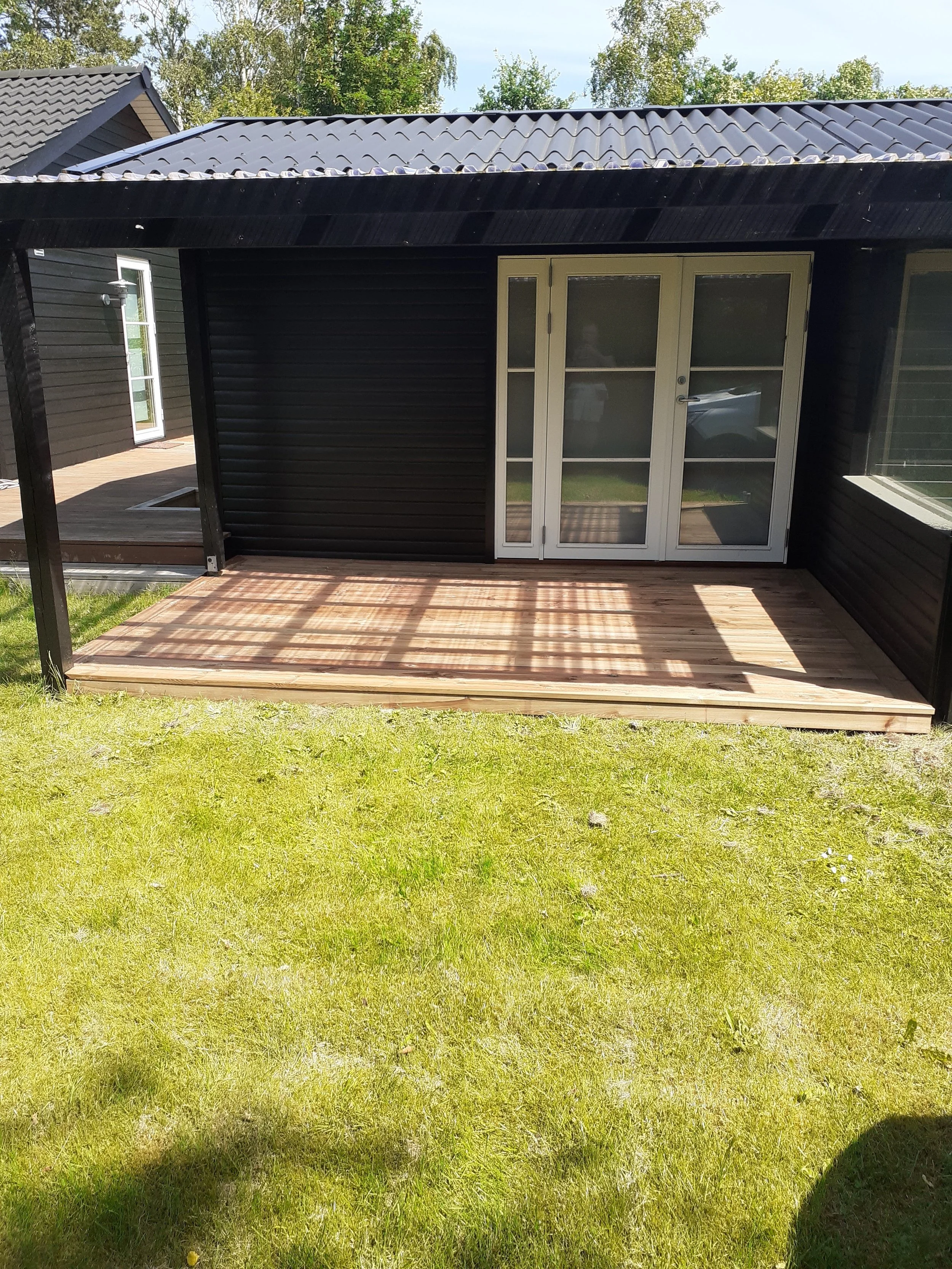 Træterrasse ved moderne bolig med store glaspartier, omgivet af græs og lignende huse i området.