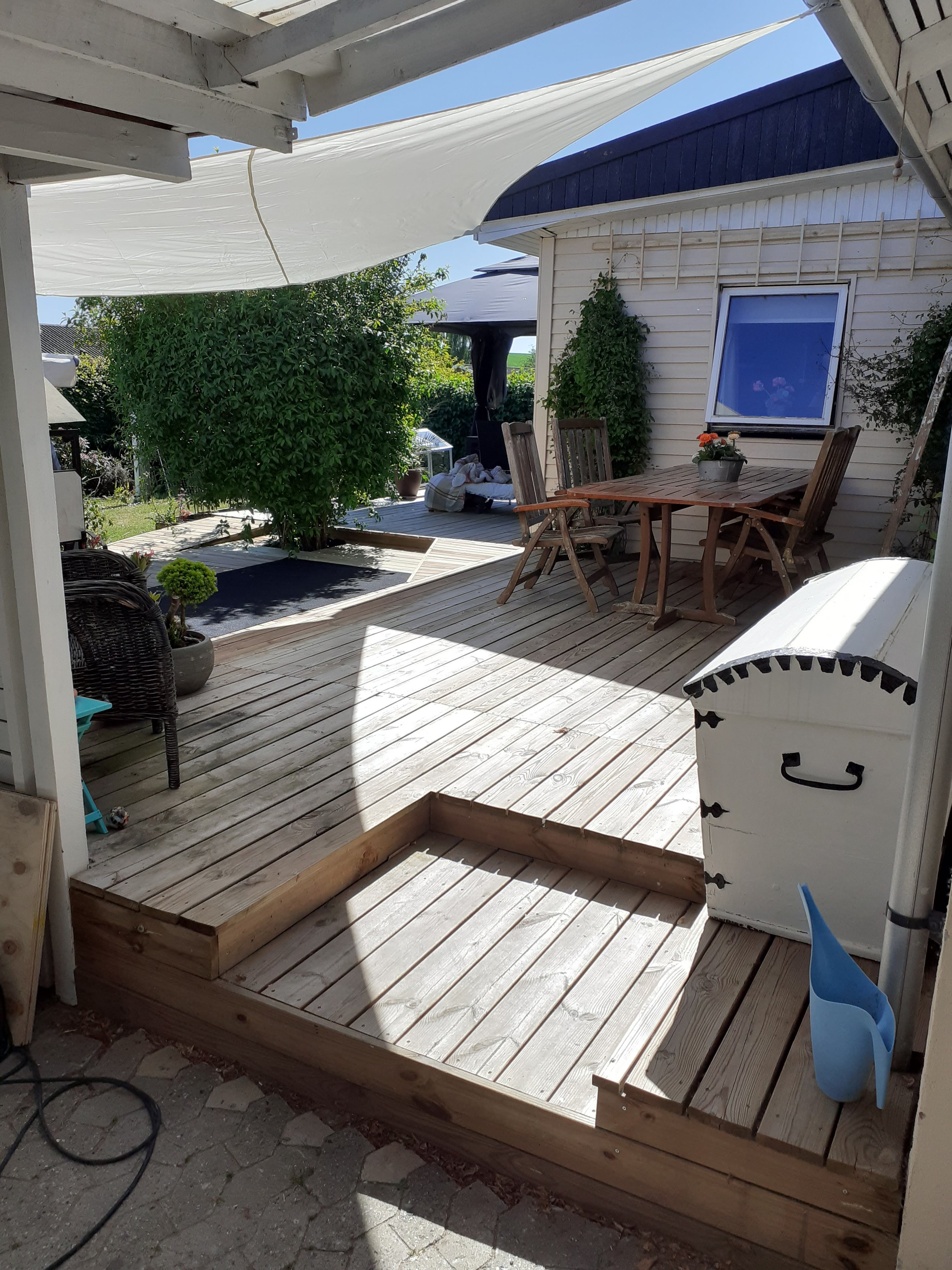 Nybygget træterrasse med havemøbler og et hvidt skur, omgivet af træer og buske.