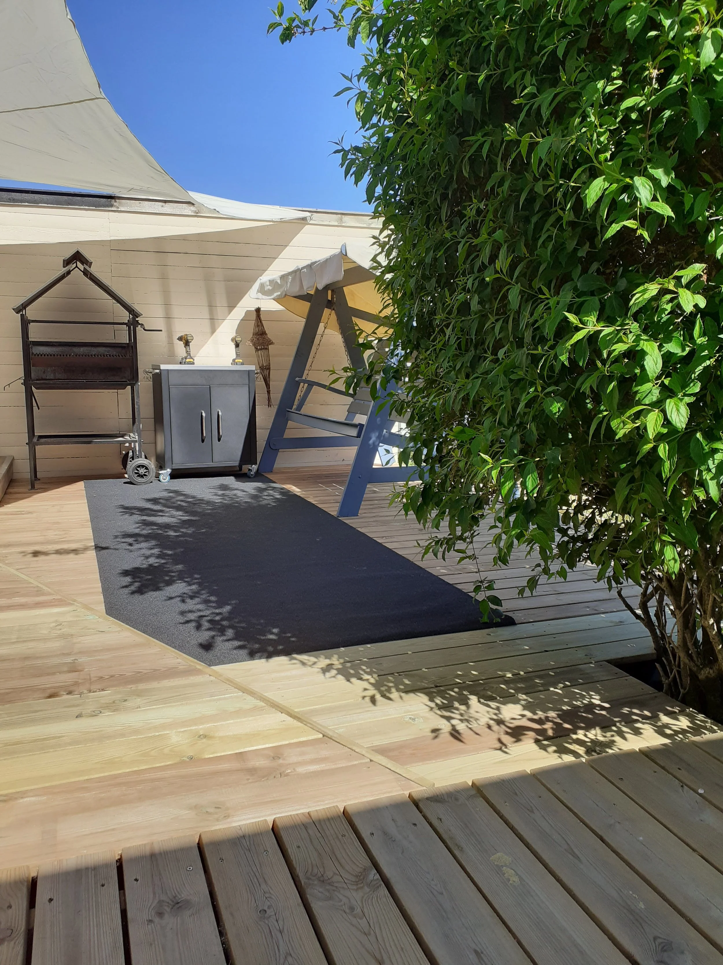 Terrassemiljø med tæppe, grill, opbevaringsskab og hængebænk under åben himmel, omgivet af grøn beplantning.