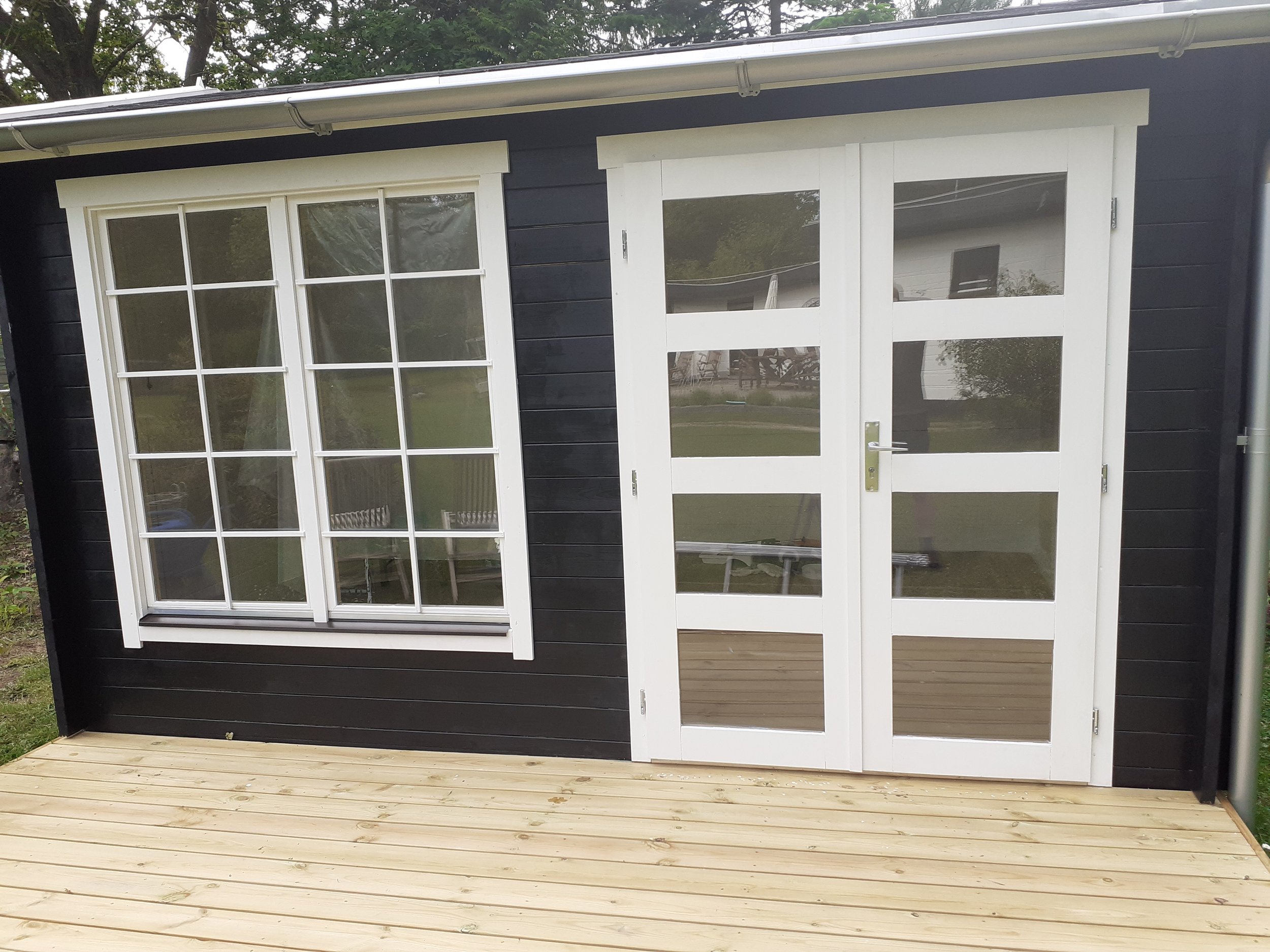 Sort træbygning med stor terrasse, store vinduer og dobbeltdør med glaspartier.