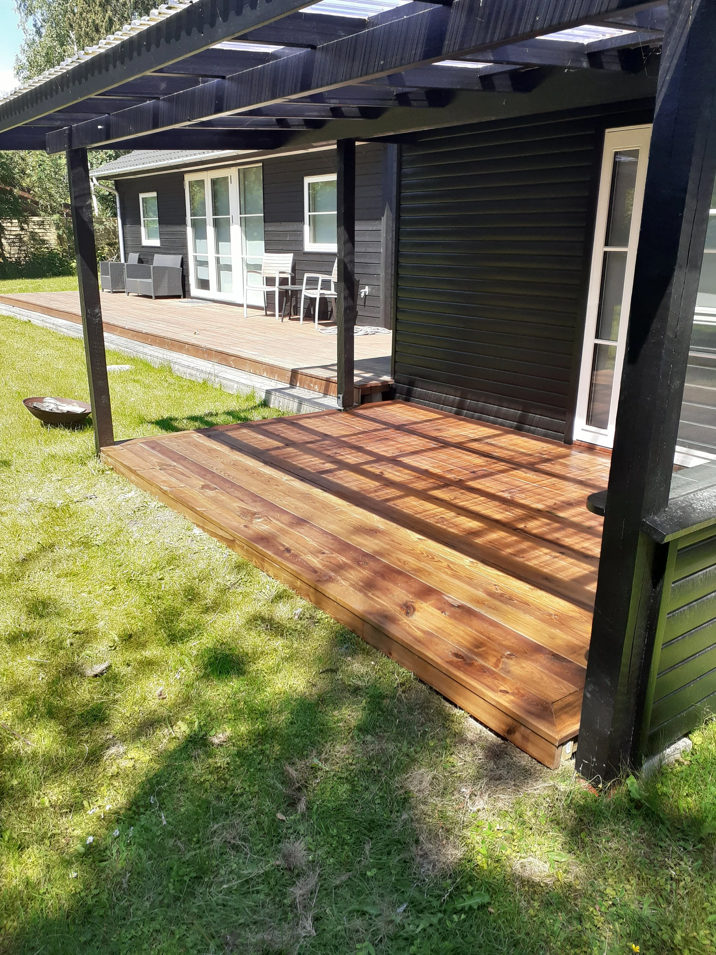 Træterrasse ved sort hus med hvide glaspartier, overdækket af en pergola og med havemøbler i baggrunden.