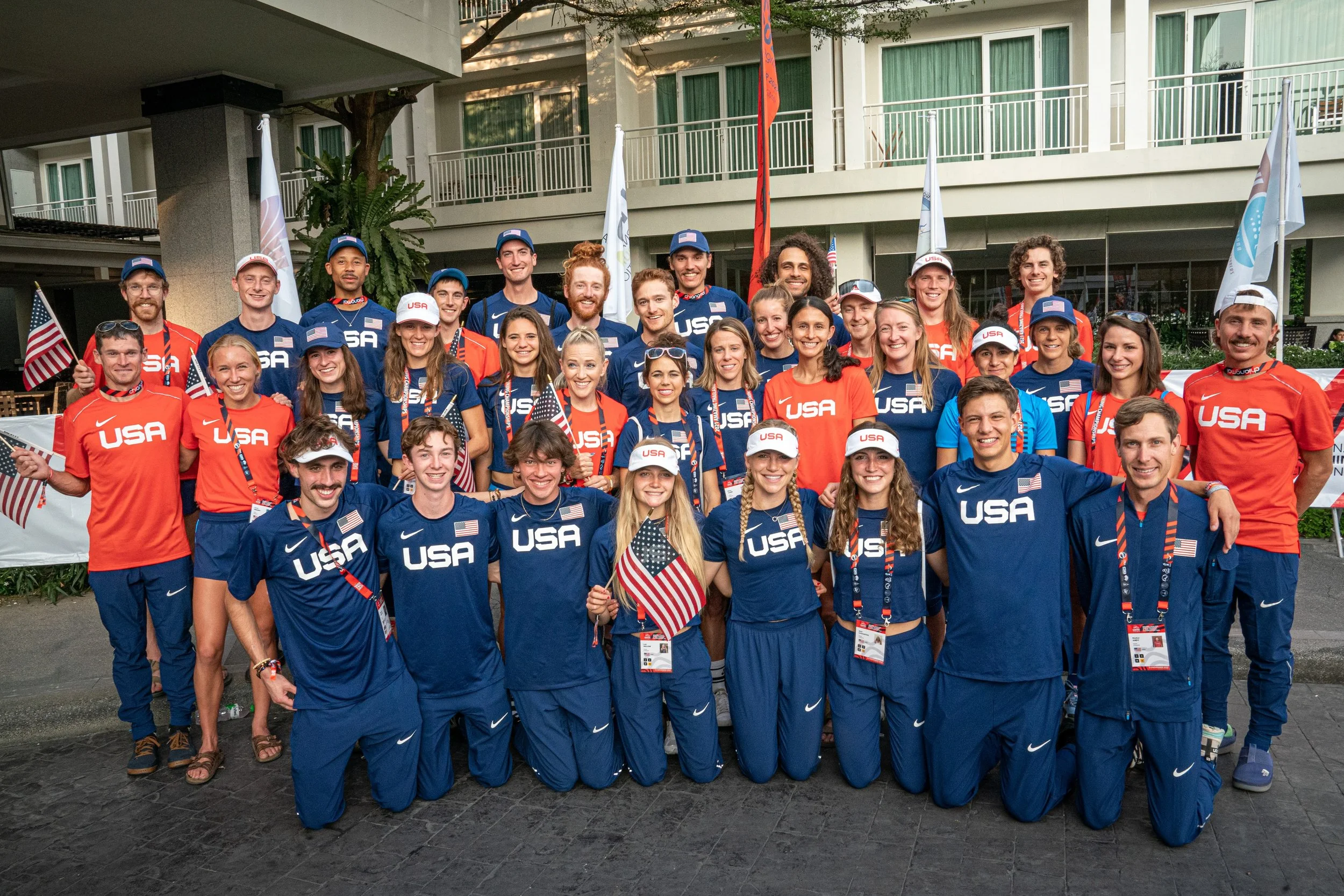 A large group of athletes, coaches, and officials from Team USA, smiling and posing for a group photo outdoors in front of a hotel or building. They are wearing red, white, and blue sportswear with "USA" and American flags, some holding small American flags.