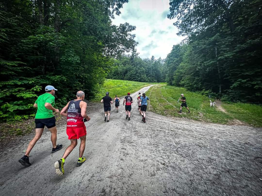 People running on a dirt trail in a forested area, with trees on both sides and a grassy hill ahead.
