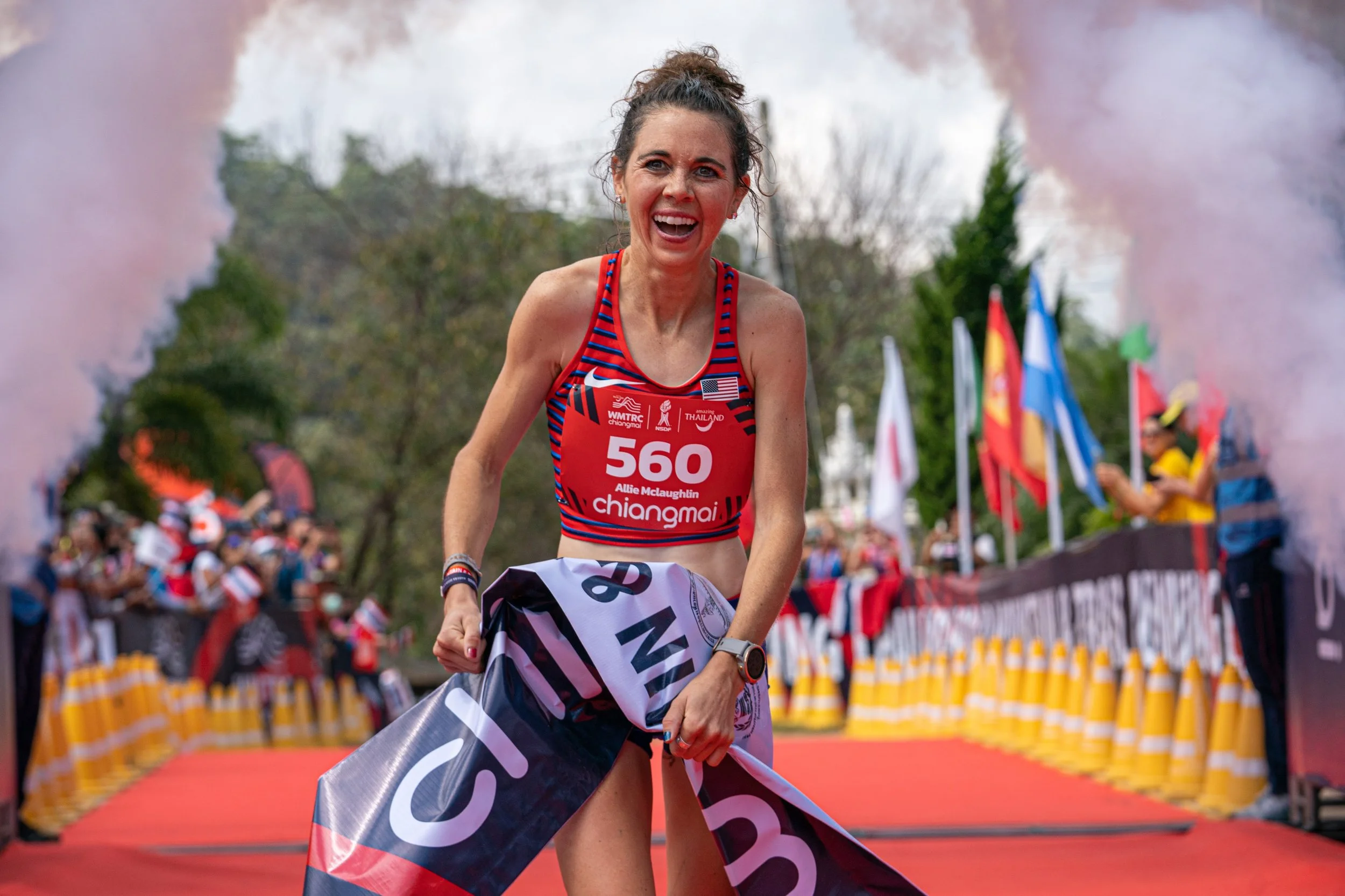 A female runner in a red and blue striped sports bra celebrating as she crosses the finish line of a race, holding a banner with a crowd cheering in the background.