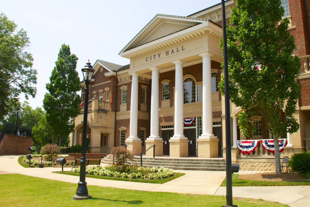 Front view of a city hall building decorated with red, white, and blue bunting for a patriotic celebration, surrounded by trees, lampposts, and a well-maintained lawn.